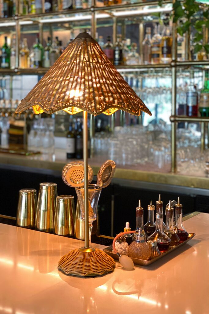 Close-up of a rattan table lamp on the Pink Cabana bar in Palm Springs, surrounded by brass cocktail shakers, bar tools, and vintage-style glass bitters bottles.