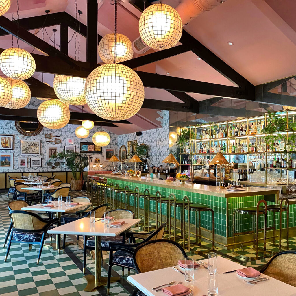 Interior of the Pink Cabana restaurant in Palm Springs featuring a green tile bar, rattan lamps, globe pendant lights, tropical wallpaper, and modern aesthetic dining tables.