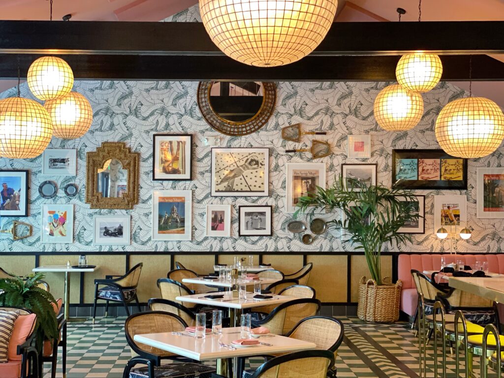 Interior of the Pink Cabana restaurant at the Sands Hotel in Palm Springs, featuring tropical wallpaper, gallery wall art, rattan chairs, pink banquettes, globe pendant lights, and vintage-inspired decor.