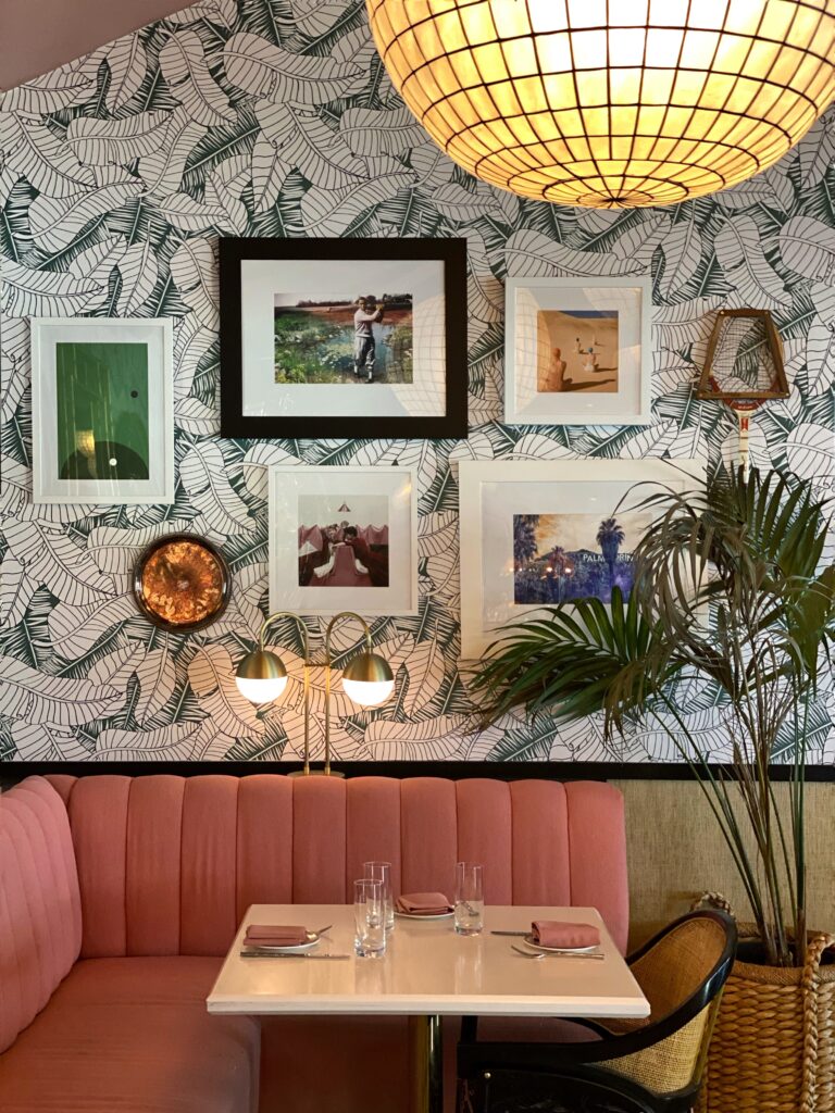 Aesthetic booth seating at the Pink Cabana in Palm Springs, featuring a blush pink banquette booth seating, tropical leaf wallpaper, gallery wall art, brass table lamps, and rattan accents.