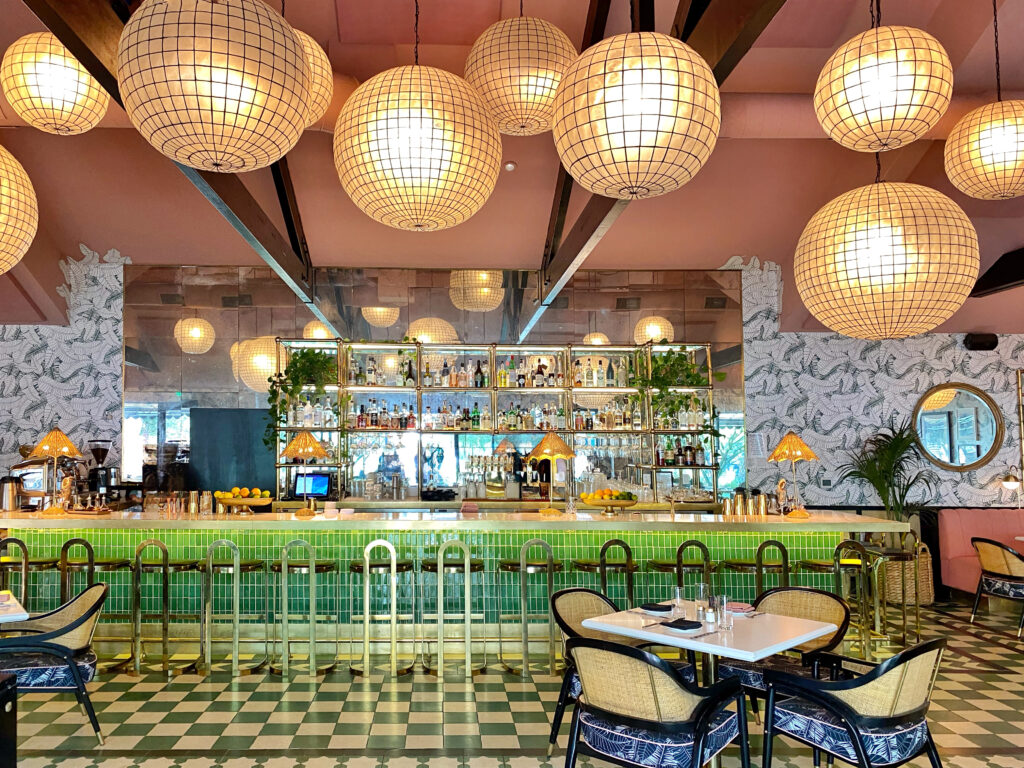 The Pink Cabana restaurant at The Sands Hotel in Indian Wells, featuring a green tile bar with brass accents, oversized globe pendant lights, patterned wallpaper, and cane-back chairs in a chic desert-Mediterranean design.