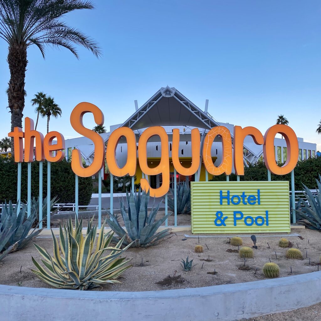 Bright orange ‘The Saguaro’ sign in front of the hotel’s entrance in Palm Springs, surrounded by desert landscaping with agave and small cacti under a clear blue sky.