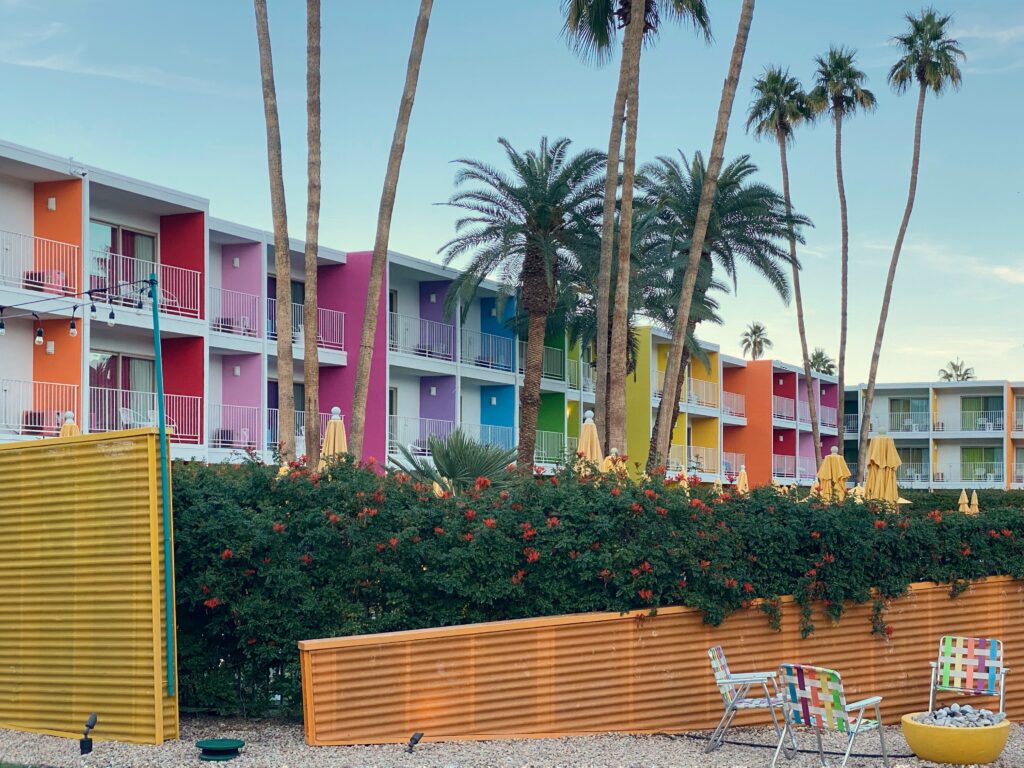 Palm Springs Aesthetic Travel Guide - The Saguaro Palm Springs featuring its signature rainbow-colored balconies, tall palm trees, and a desert courtyard with corrugated yellow panels and colorful woven chairs.