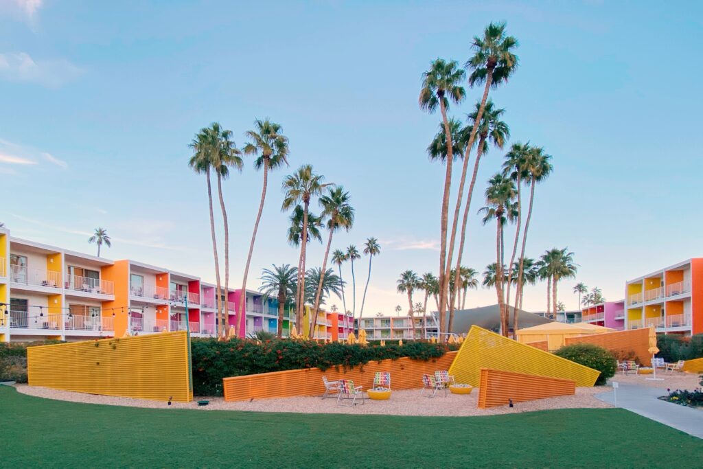 The Saguaro Palm Springs with its iconic rainbow-colored balconies, tall palm trees, and playful desert-modern courtyard seating set against a clear blue sky.