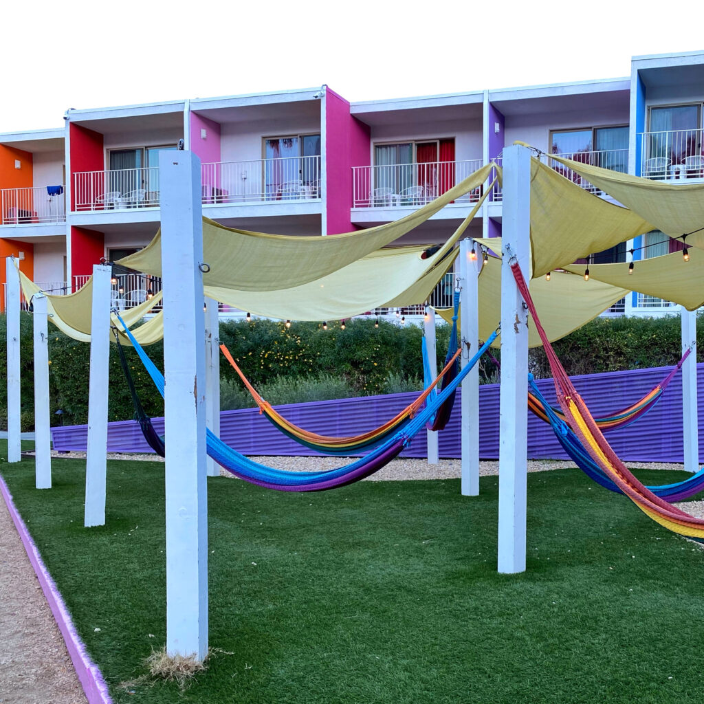 Colorful hammock garden at The Saguaro Palm Springs with rainbow woven hammocks, shade canopies, string lights, and the hotel’s vibrant pink, orange, purple, and blue balconies in the background.
