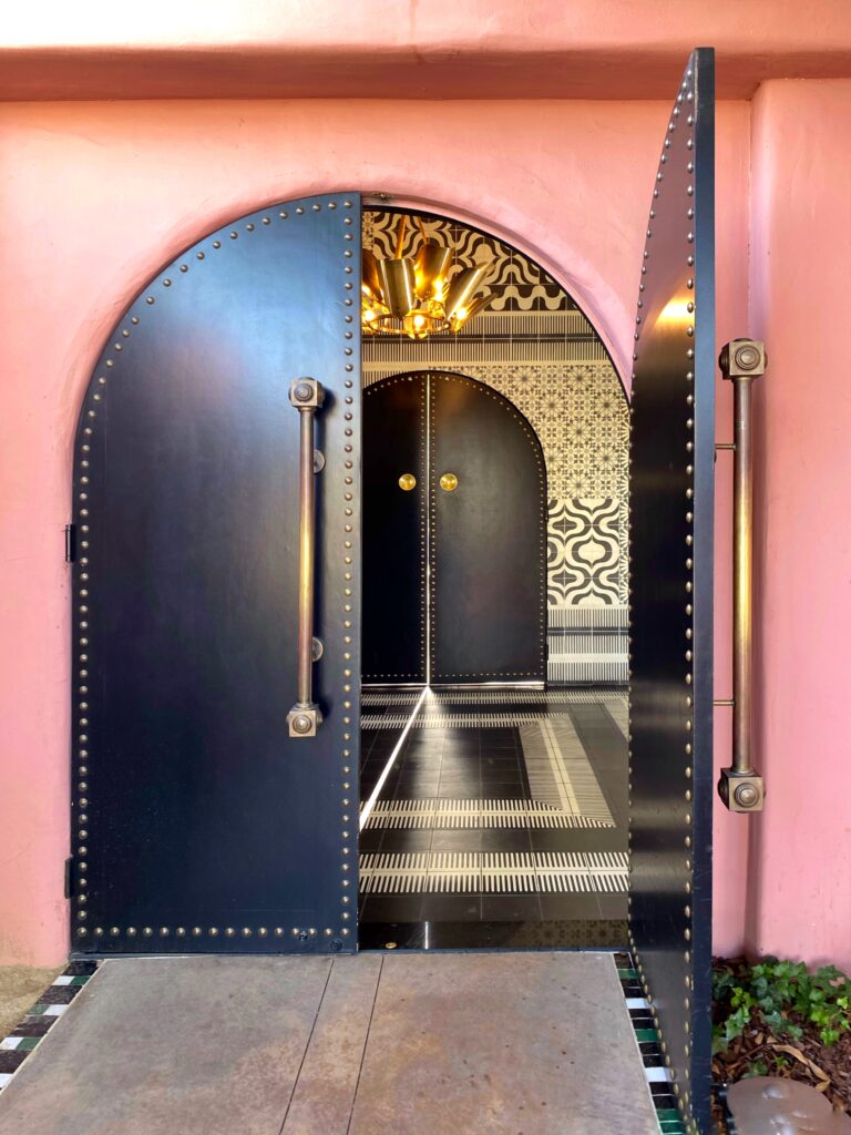 Close-up of the black arched entry doors at The Sands Hotel in Indian Wells, featuring brass hardware, pink stucco walls, and a view into a patterned Moroccan-style tile interior with warm gold lighting — a signature Palm Springs boutique-hotel aesthetic.