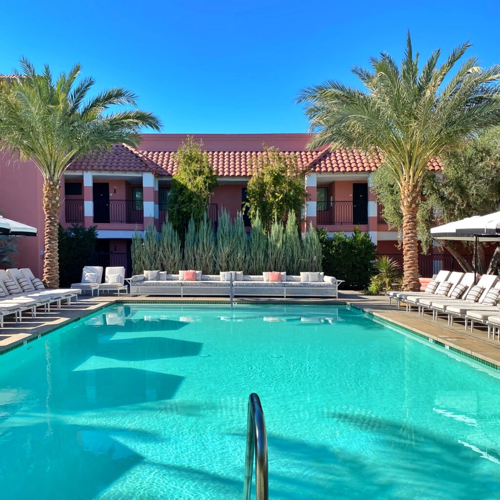 Poolside scene at The Sands Hotel in Indian Wells featuring turquoise water, striped loungers, tall palm trees, and the hotel’s signature pink Mediterranean-inspired architecture under a clear blue sky.