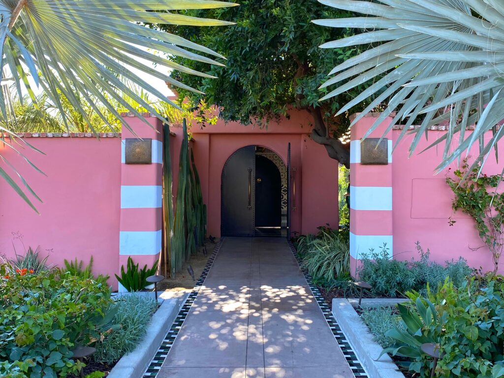Palm Springs–style entrance at The Sands Hotel in Indian Wells, featuring pink stucco walls with white stripes, tall desert cacti, lush landscaping, and a black arched Moroccan-inspired doorway framed by palm fronds.