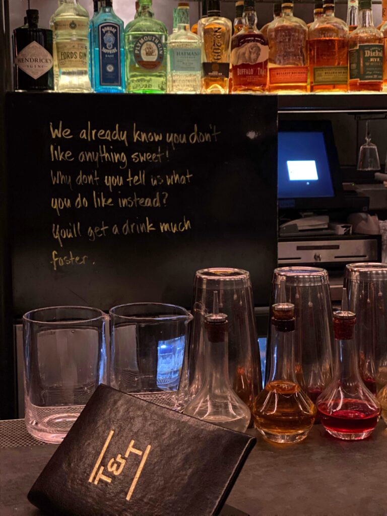 Close-up bar detail at Truss & Twine in Palm Springs with bottles of gin and whiskey on a backlit shelf, glassware and bitters bottles on the counter, and a sassy handwritten note on the chalkboard about drink preferences.