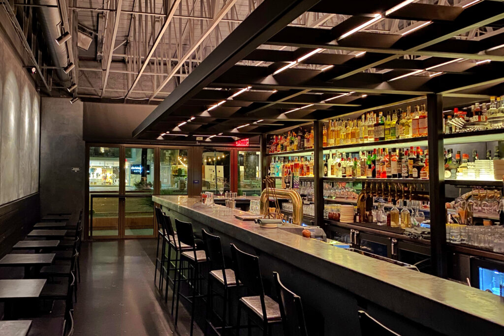 Industrial-style interior of Truss & Twine in Palm Springs with a long concrete bar, black metal shelving lined with spirits, warm backlighting, and exposed ceiling trusses for a modern desert cocktail atmosphere.
