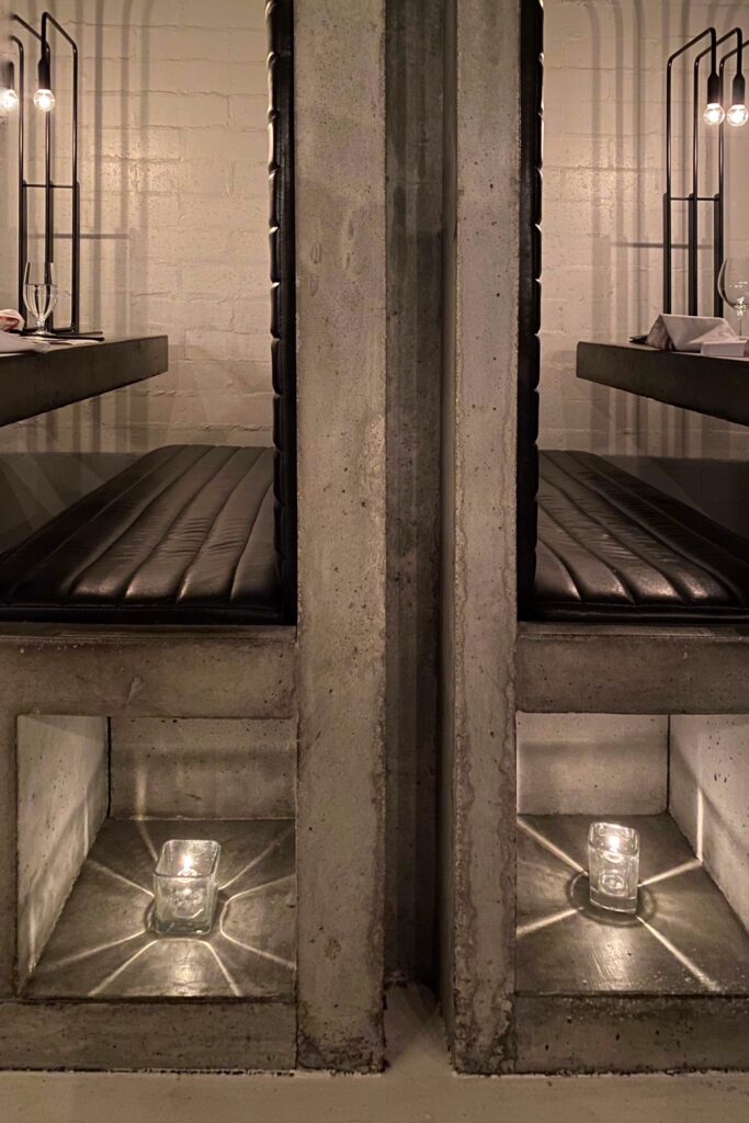 Concrete booth detail at Workshop Kitchen + Bar in Palm Springs, showing black leather bench seating, minimalist pendant lighting, and candlelit niches that cast star-shaped reflections.
