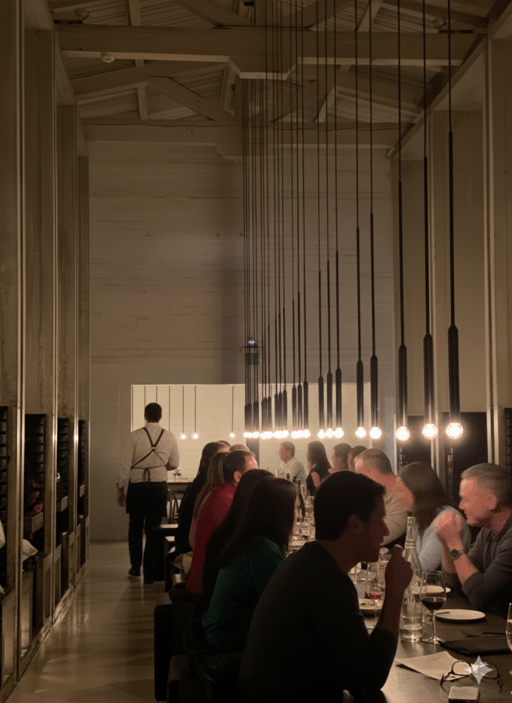 Dimly lit industrial dining room at Workshop Kitchen + Bar in Palm Springs, featuring dramatic rows of hanging pendant bulbs, tall concrete partitions, and guests seated along a long communal table.