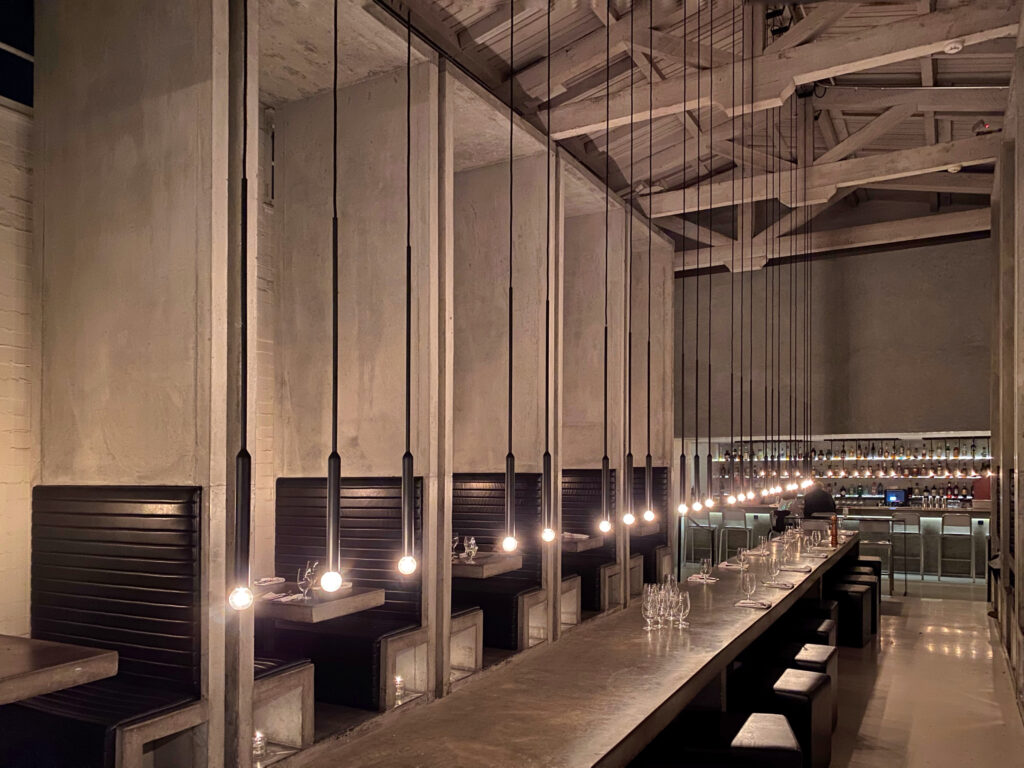 Industrial-chic interior of Workshop Kitchen + Bar in Palm Springs, featuring dramatic concrete architecture, a central long communal table, leather booths, and minimalist pendant lighting — a sleek design destination for modern dining and cocktails in the desert.