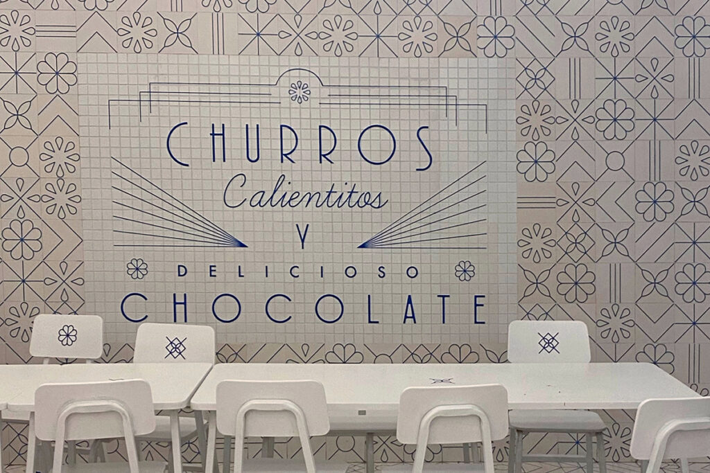 Blue-and-white tiled wall inside El Moro Churrería in Mexico City, featuring geometric patterns and typography for churros and chocolate, paired with simple white café tables and chairs.