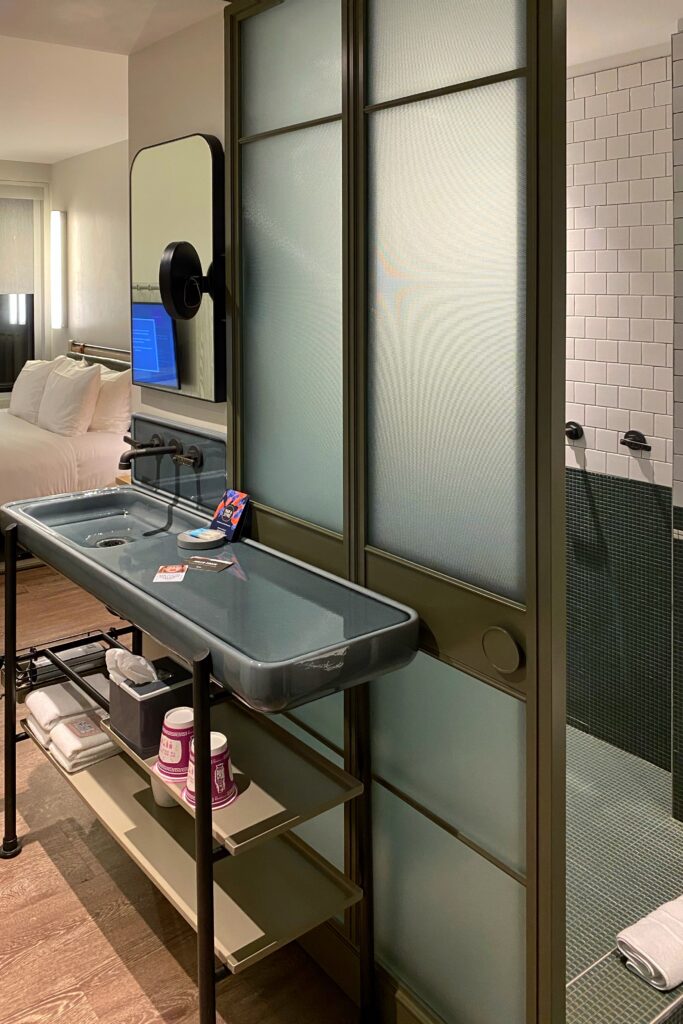 Compact modern hotel bathroom at Moxy Times Square featuring a sleek wall-mounted sink with open shelving, frosted glass sliding doors, matte black fixtures, and a walk-in tiled shower—an efficient design solution for small urban spaces.