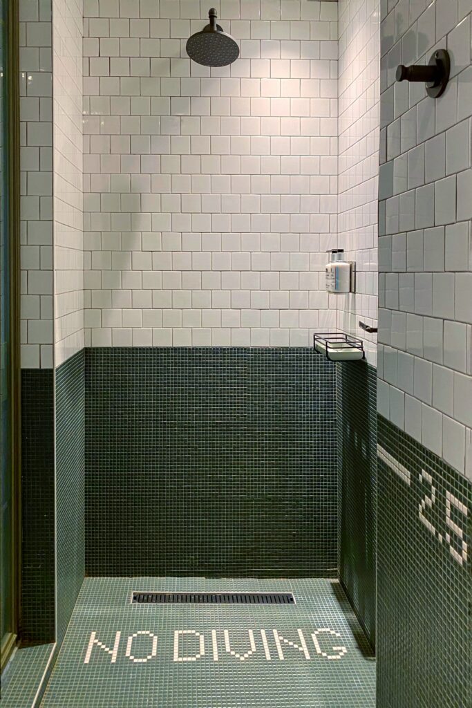 Playful hotel shower design at Moxy Times Square featuring green mosaic tile flooring with "NO DIVING" typography, matte black rain showerhead, and white subway tile walls—blending humor with urban modern style in a compact space.