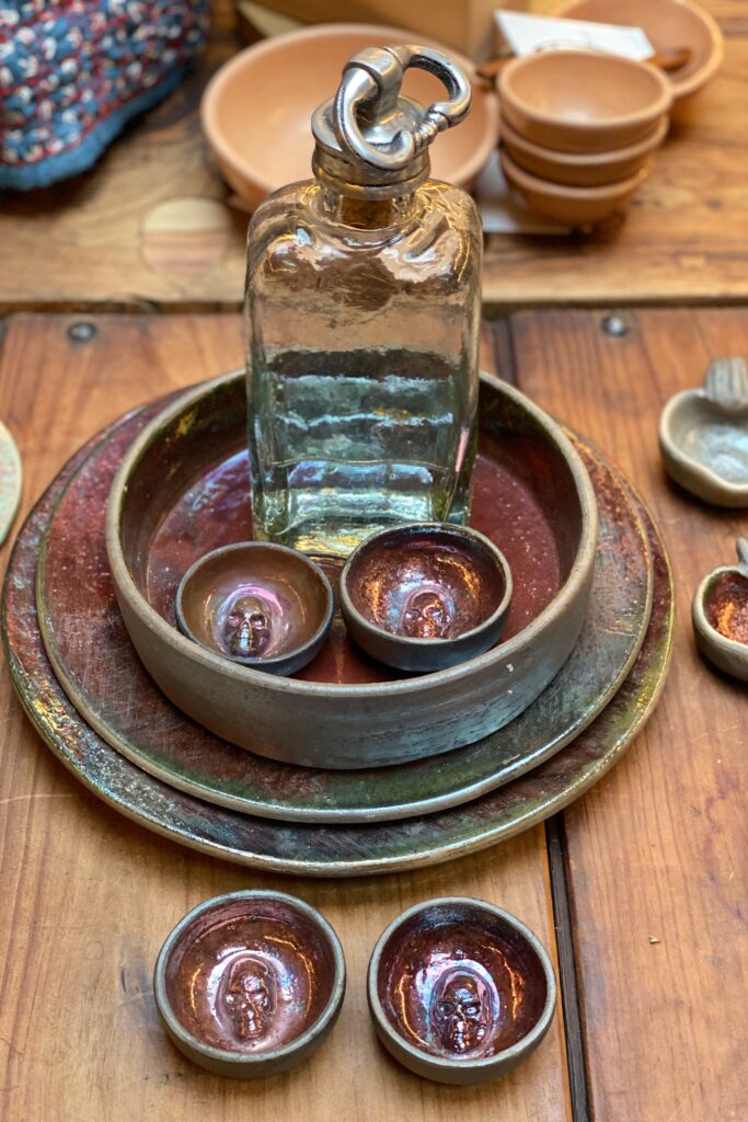 Artisan ceramic dishes with subtle skull details displayed inside the Tetetlán shop in Mexico City, layered with a handblown glass bottle on a rustic wood table; the earthy iridescent glazes, sculptural forms, and quiet craftsmanship reflect Tetetlán’s thoughtful blend of contemporary design, Mexican tradition, and curated travel retail.