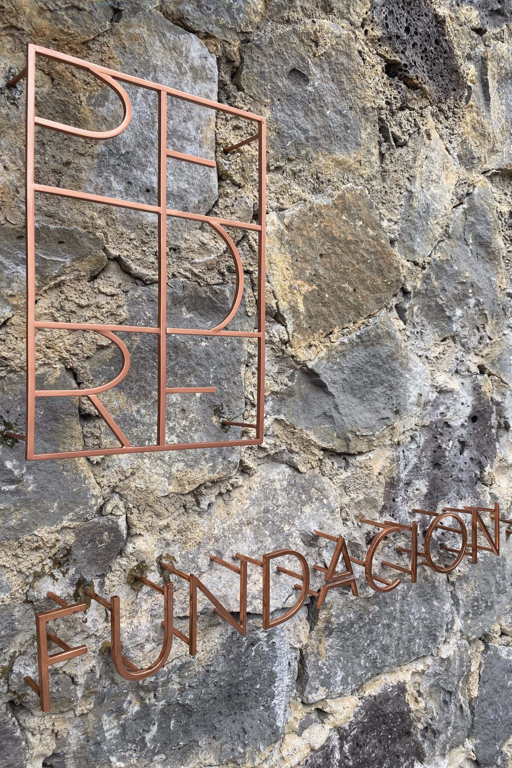 opper-toned metal signage for Fundación Tetetlán mounted on a rugged volcanic stone wall in Mexico City; the minimalist typography and raw material palette reflect the site’s architectural identity, blending contemporary design with Mexico’s geological and cultural landscape.