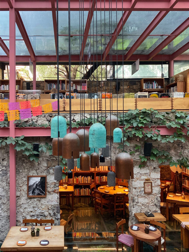 Interior view of Tetetlán in Mexico City, featuring a double-height dining space with suspended sculptural pendant lights, exposed stone walls, indoor greenery, and a mezzanine library; contemporary Mexican architecture blending brutalist structure with warm wood and artisanal details.