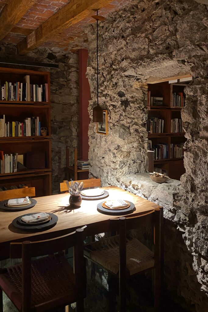 Intimate dining room inside Tetetlán in Mexico City featuring a solid wood table set beneath exposed volcanic stone walls and brick ceilings, flanked by built-in bookshelves; warm, focused lighting and handcrafted tableware create a library-like atmosphere that reflects Tetetlán’s thoughtful fusion of architecture, dining, and cultural experience.