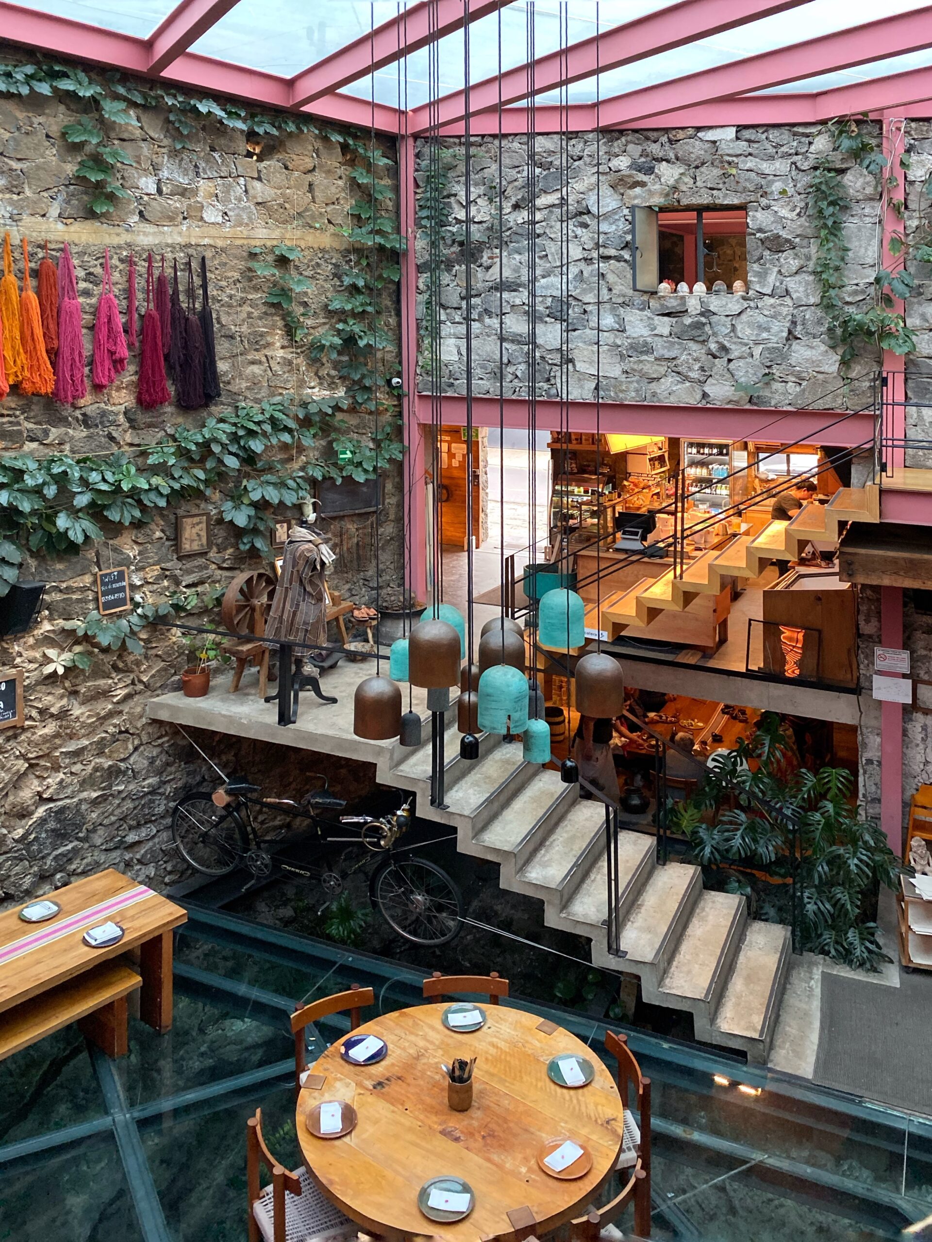 Multi-level interior of Tetetlán restaurant in Mexico City featuring exposed volcanic stone walls, a glass floor dining area, and sculptural concrete stairs accented by hanging ceramic pendant lights; pink steel beams, trailing greenery, and warm wood tables highlight Tetetlán’s iconic architecture and its immersive blend of modern Mexican design, dining, and travel experience.