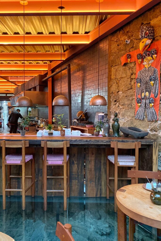 Open kitchen bar inside Tetetlán restaurant in Mexico City featuring a wood counter set above a glass floor, sculptural pendant lights, and exposed volcanic stone walls; warm orange ceiling beams, handcrafted stools, and contemporary Mexican artwork create a richly atmospheric dining space that blends architecture, craft, and modern design.