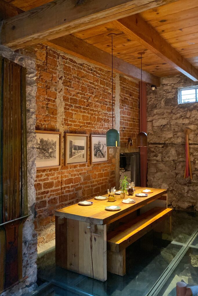 Private dining room at Tetetlán in Mexico City featuring a long solid-wood table set beneath exposed brick and volcanic stone walls, with warm timber ceiling beams and minimalist pendant lighting; framed black-and-white photography, handcrafted tableware, and a glass floor create an intimate, design-led dining space rooted in material honesty and contemporary Mexican architecture.