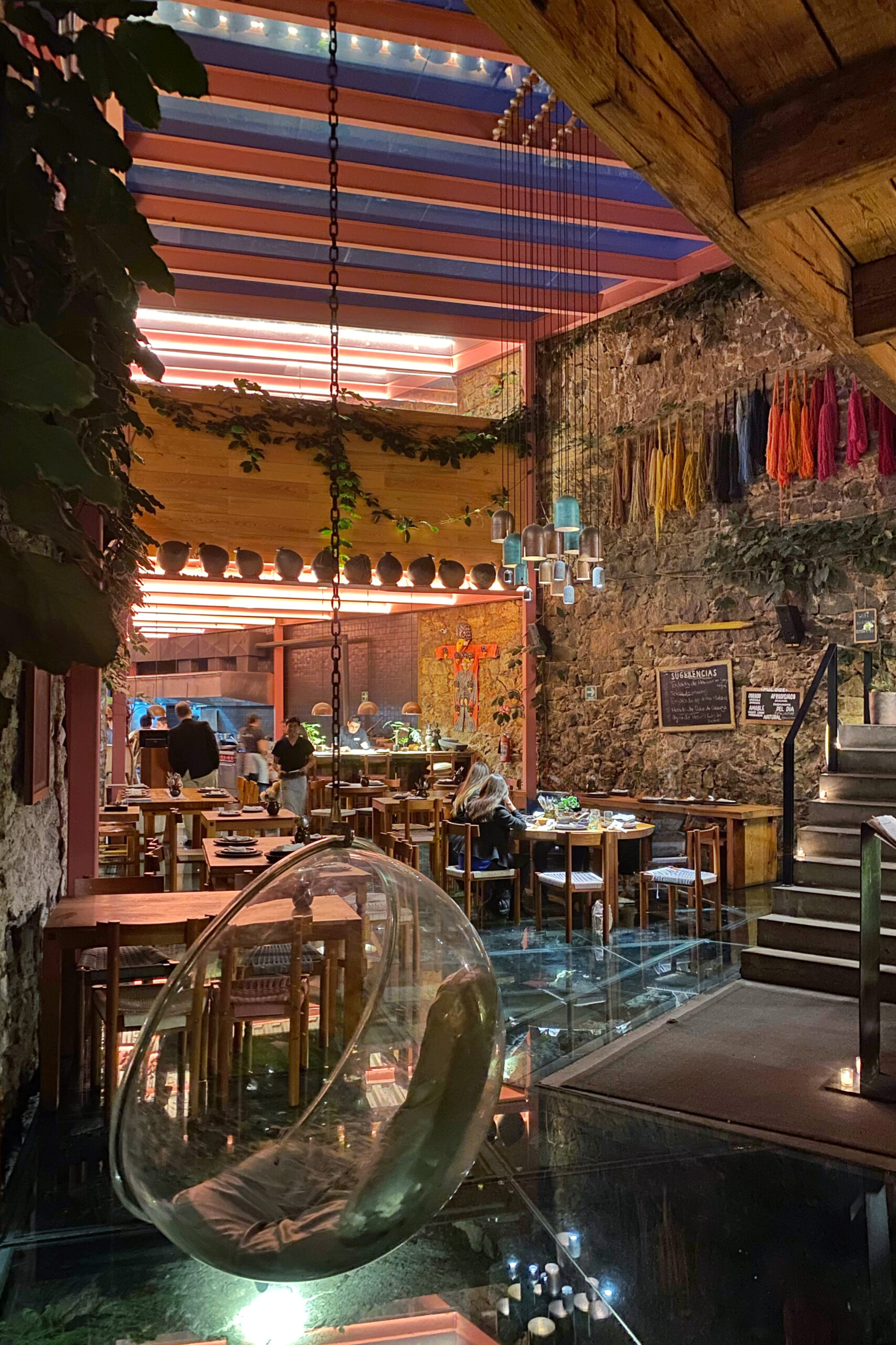 Evening view inside Tetetlán restaurant in Mexico City featuring volcanic stone walls, a glass floor illuminated with candlelight, and a dramatic open dining room framed by wood tables, a swing bubble chair, and hanging sculptural pendants; pink steel beams, colorful textile accents, and warm ambient lighting create a richly layered, design-driven dining experience rooted in contemporary Mexican architecture.