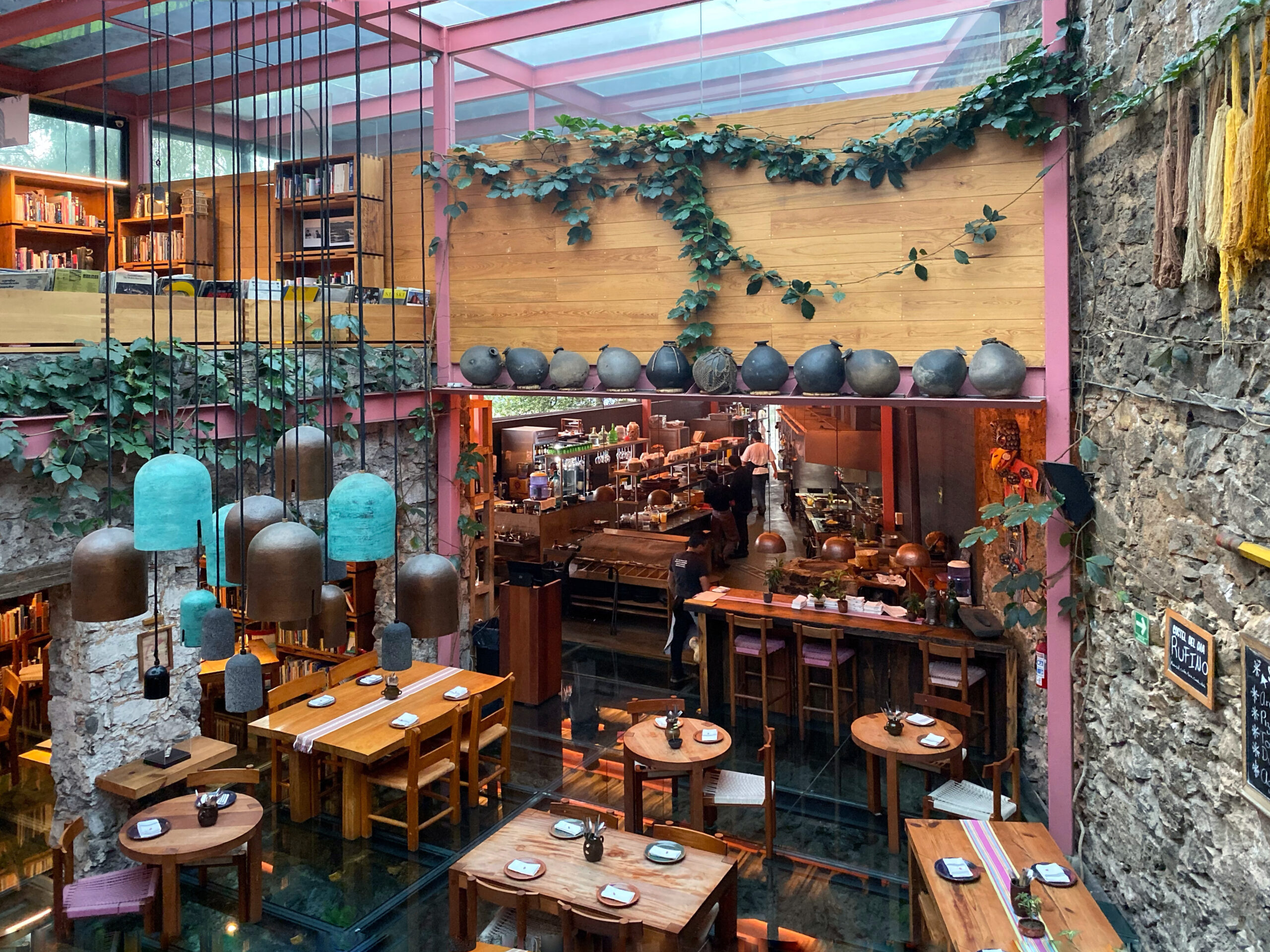 Interior view of Tetetlán restaurant in Mexico City showcasing volcanic stone walls, a glass floor, and a dramatic multi-level dining space framed by pink steel beams, hanging sculptural lights, and lush greenery; warm wood tables, an open kitchen, and layered architectural details reflect Tetetlán’s celebrated blend of modern Mexican design, hospitality, and immersive travel dining.