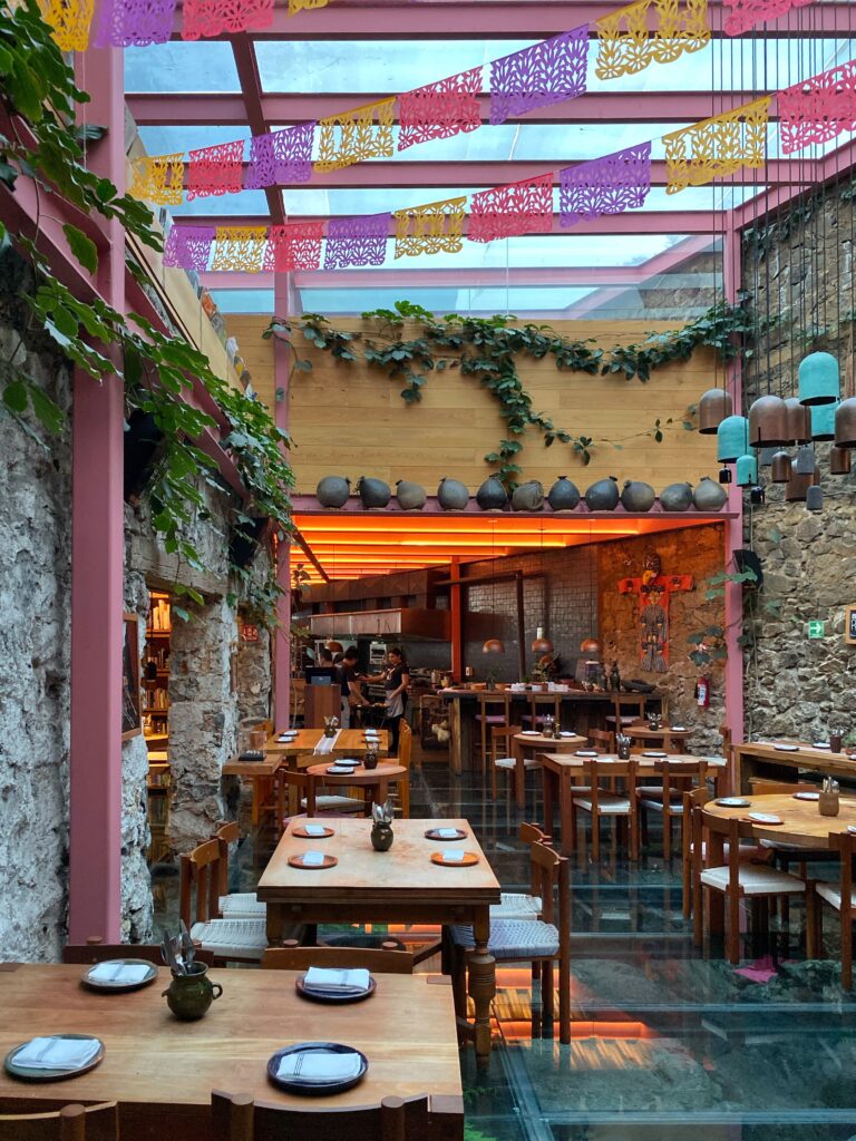 Daytime interior of Tetetlán restaurant in Mexico City featuring a glass ceiling framed by pink steel beams, colorful papel picado banners, and exposed volcanic stone walls; warm wood tables, a glowing open kitchen, trailing greenery, and sculptural lighting create a vibrant yet refined dining space that blends contemporary Mexican design with cultural tradition.