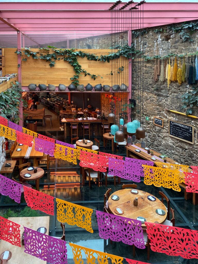 Overhead view of Tetetlán restaurant in Mexico City featuring a dramatic glass floor dining area, exposed volcanic stone walls, and pink steel beams beneath a skylit ceiling; colorful papel picado banners, trailing greenery, sculptural pendant lights, and warm wood tables create a vibrant yet architecturally refined dining space rooted in contemporary Mexican design.