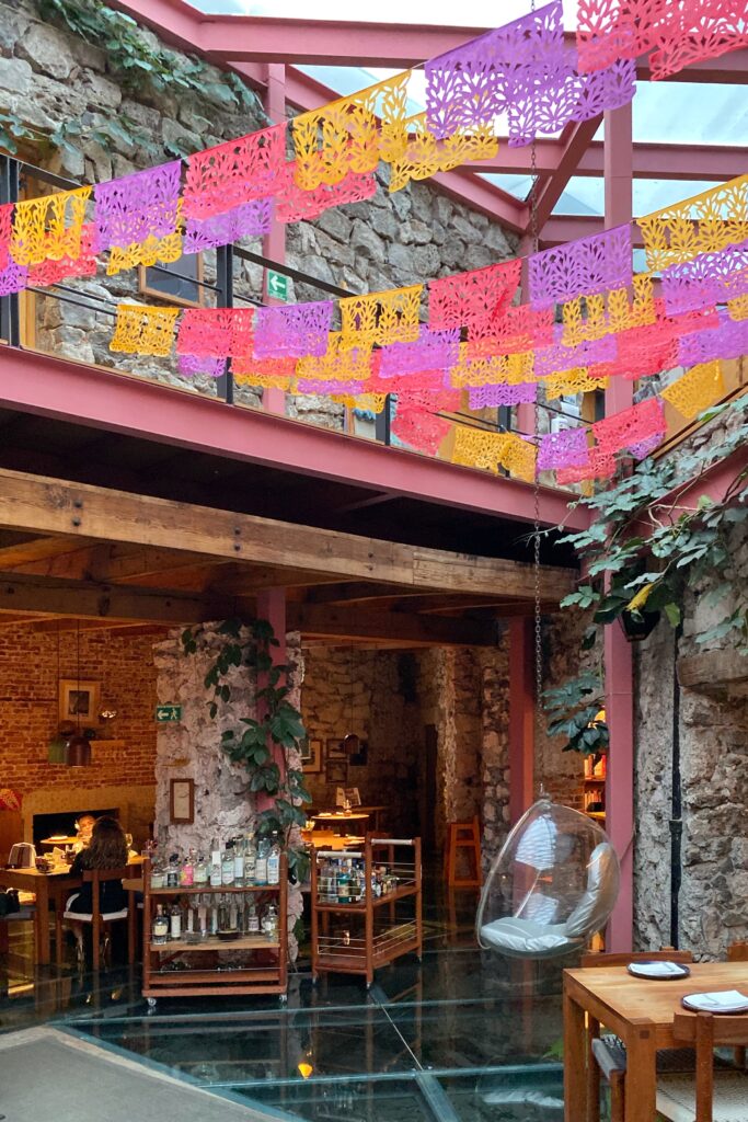 Interior courtyard at Tetetlán in Mexico City featuring colorful papel picado banners strung beneath pink steel beams, exposed volcanic stone walls, and trailing greenery; a glass floor, warm wood tables, and a suspended acrylic chair create a vibrant yet architecturally grounded dining space that blends contemporary Mexican design with cultural tradition.