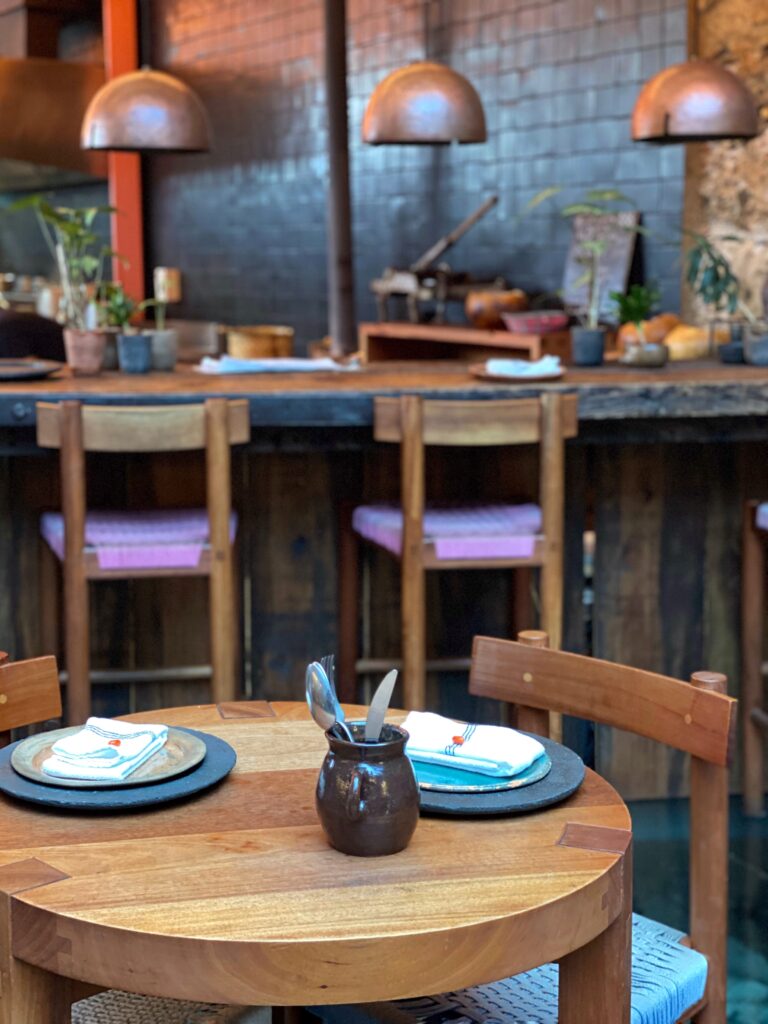 Close-up of a handcrafted wood dining table set with ceramic plates, linen napkins, and artisanal tableware inside Tetetlán restaurant in Mexico City; warm bar seating, sculptural pendant lights, and the open kitchen beyond highlight Tetetlán’s thoughtful approach to modern Mexican design, materiality, and dining experience.