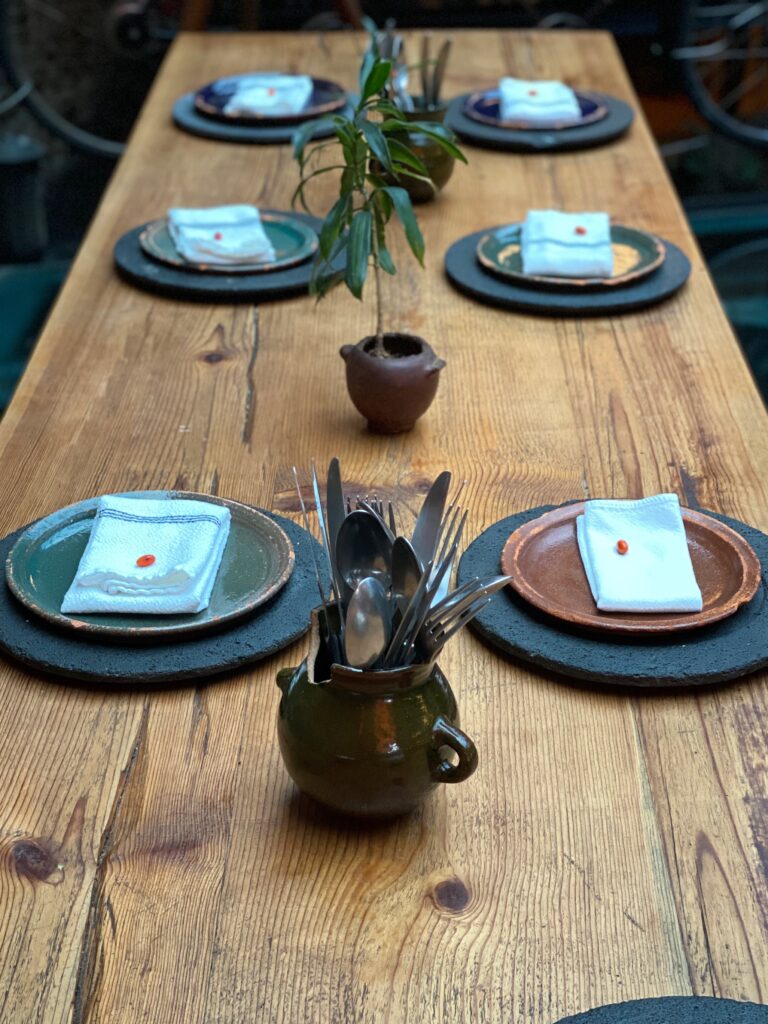 Close-up of a wooden communal dining table at Tetetlán in Mexico City, set with handcrafted ceramic plates, folded linen napkins, and artisanal cutlery held in a glazed clay vessel; minimal table styling, natural textures, and muted earth tones reflect the restaurant’s design-driven approach to contemporary Mexican dining.