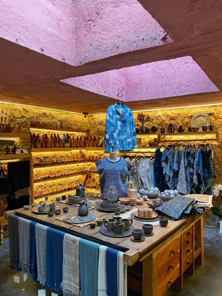 Design-forward retail interior at Tetetlán in Mexico City featuring artisan ceramics, indigo-dyed textiles, woven leather huaraches, and curated home objects displayed on a central wood table; volcanic stone walls, warm ambient lighting, and a pink concrete skylight reflect Tetetlán’s architectural heritage and its thoughtful blend of Mexican craft, travel culture, and contemporary design.