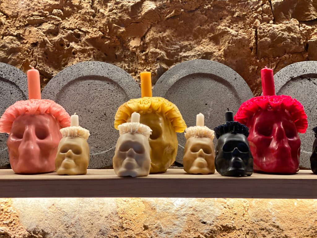 Handcrafted skull-shaped candles displayed inside the Tetetlán retail shop in Mexico City, arranged on a minimalist wooden shelf against a textured stone wall; the sculptural forms, earthy pigments, and artisanal craftsmanship reflect Tetetlán’s design-driven approach to Mexican culture, architecture, and elevated travel retail.