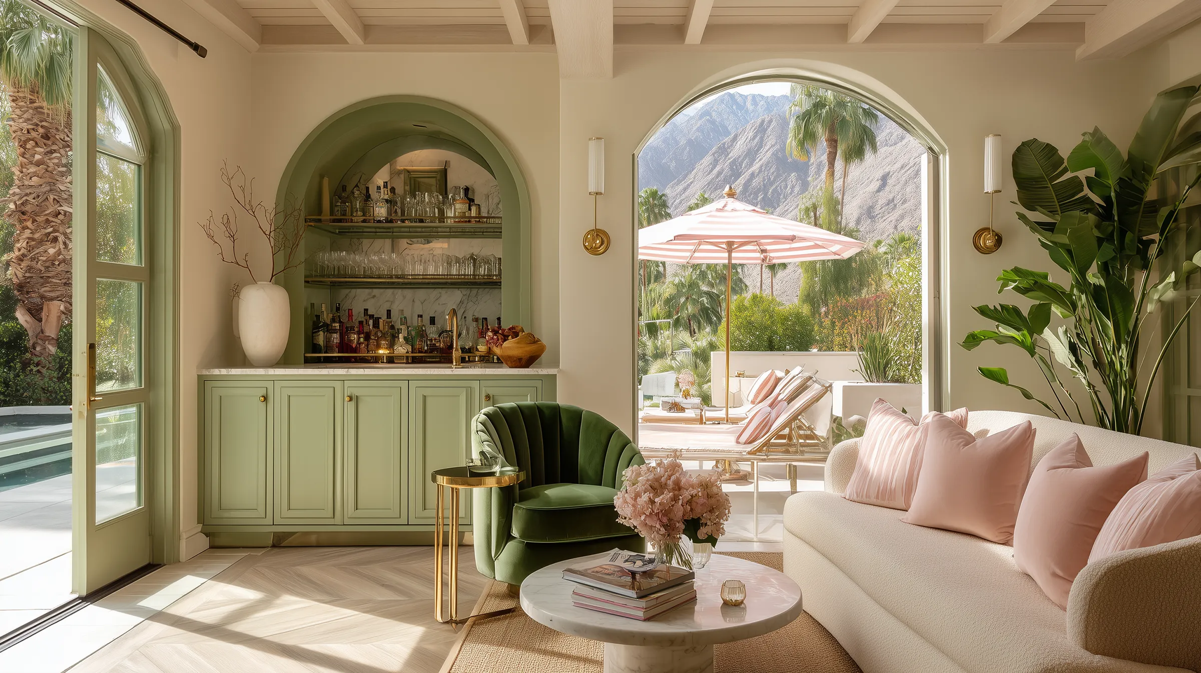 Palm Springs color palette interior with a cream bouclé sofa, blush pink throw pillows, a green velvet accent chair, sage green built-in bar with marble backsplash, brass wall sconces, and outdoor lounge chairs beneath a pink and white striped patio umbrella framed by palm trees and mountain views.
