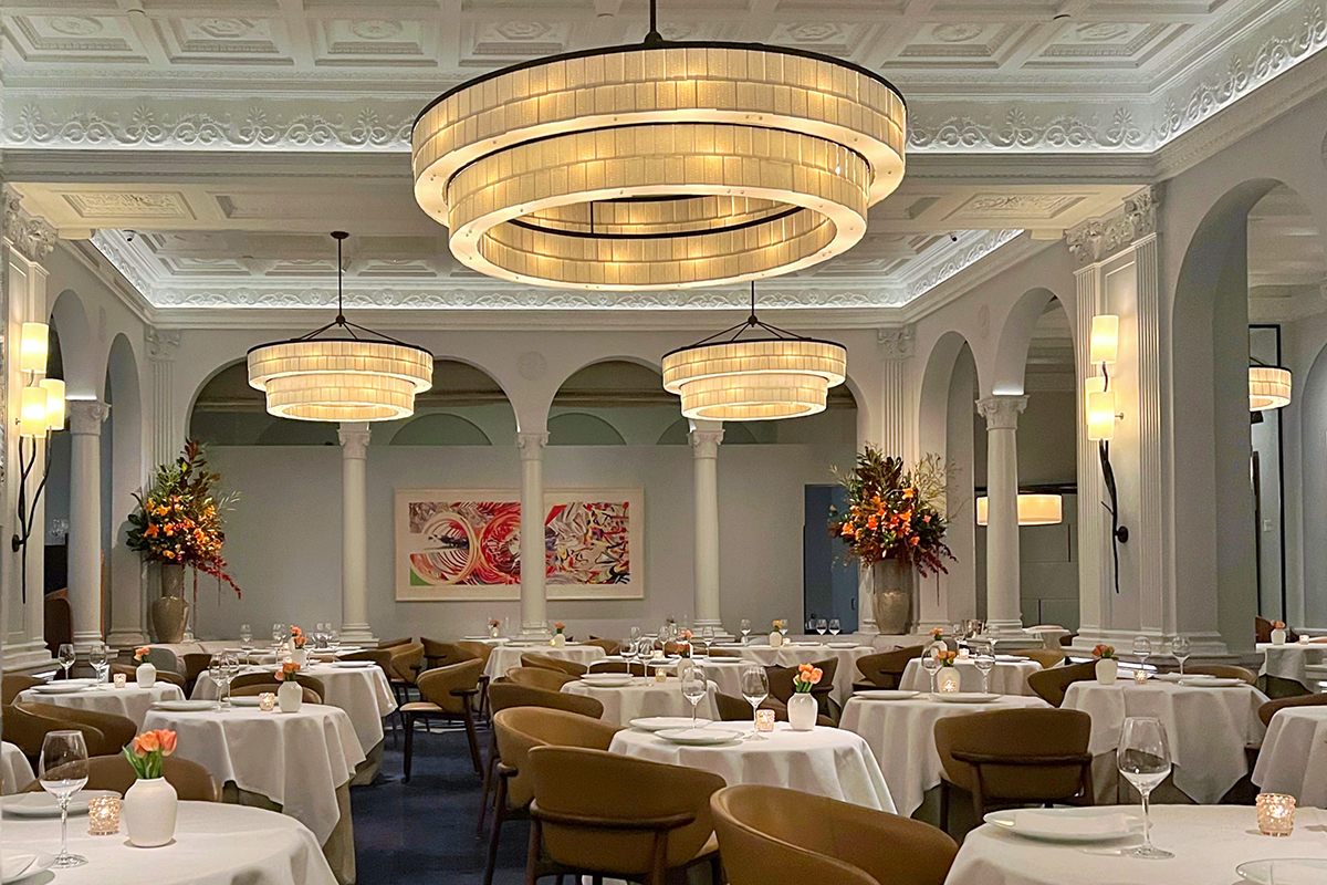 Elegant dining room at Daniel restaurant in New York City featuring grand circular tiered chandeliers, ornate coffered ceiling with plaster detailing, white arched columns, round tables with crisp white tablecloths, tan leather dining chairs, flickering candlelight, and large floral arrangements—an upscale aesthetic restaurant in NYC for design lovers and fine dining travelers.