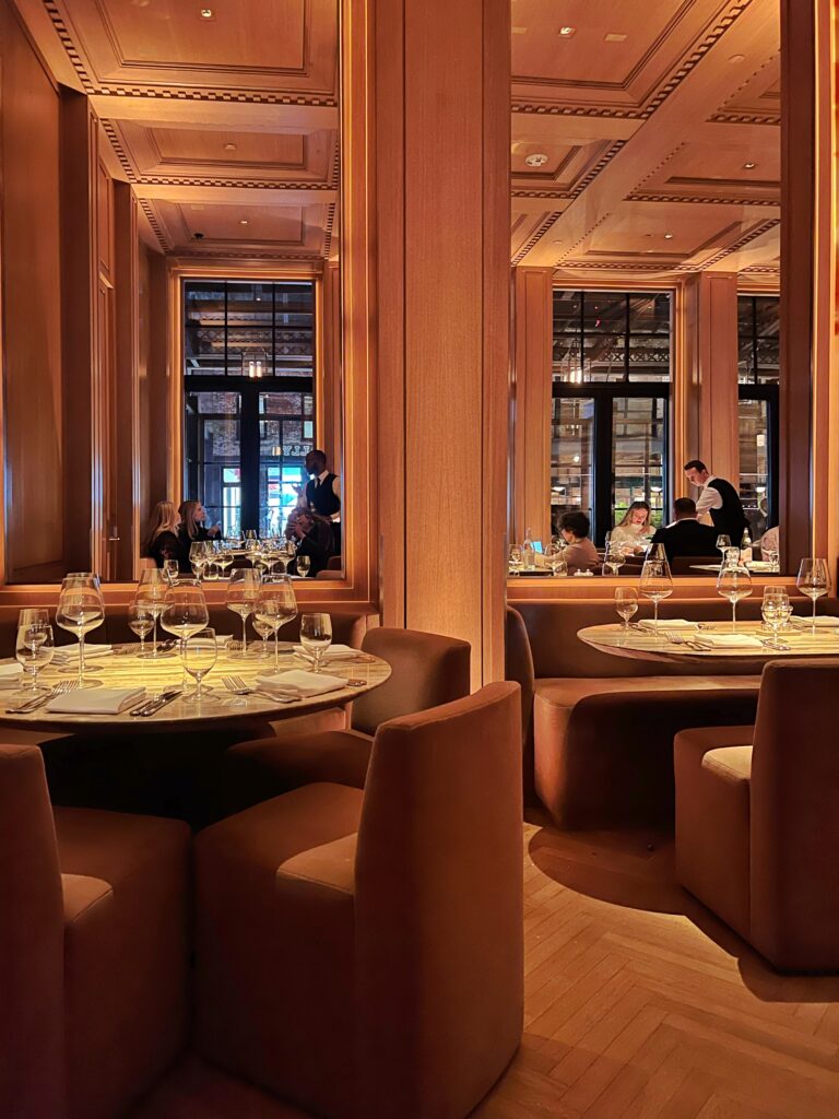 Warm wood-paneled dining room at RH Guesthouse Dining Room in New York City featuring round marble tables set with crystal stemware and linen napkins, plush caramel cube chairs and banquettes, floor-to-ceiling millwork, and coffered ceilings with soft cove lighting—an intimate, ultra-luxury aesthetic restaurant in NYC for interior design lovers and sophisticated travelers.