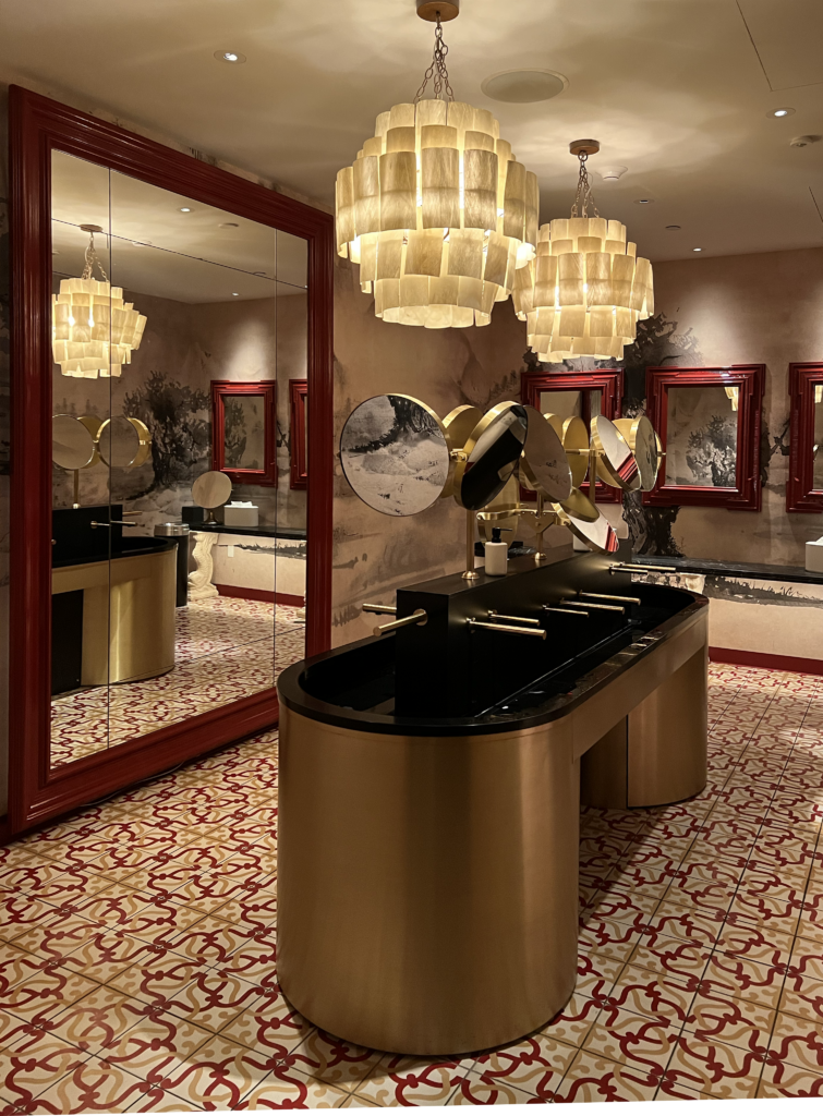 Powder room at The Ritz-Carlton New York, NoMad with patterned red-and-cream tile flooring, oxblood-painted mirror frames, tiered alabaster chandeliers, a two-sided brushed brass vanity, and mural-style walls.