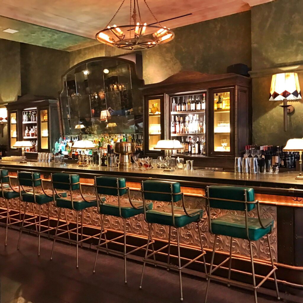 Bar at The Beekman, New York with green leather stools, carved metal bar detailing, antique mirror panels, dark wood liquor cabinets, and moody hand-painted green walls.