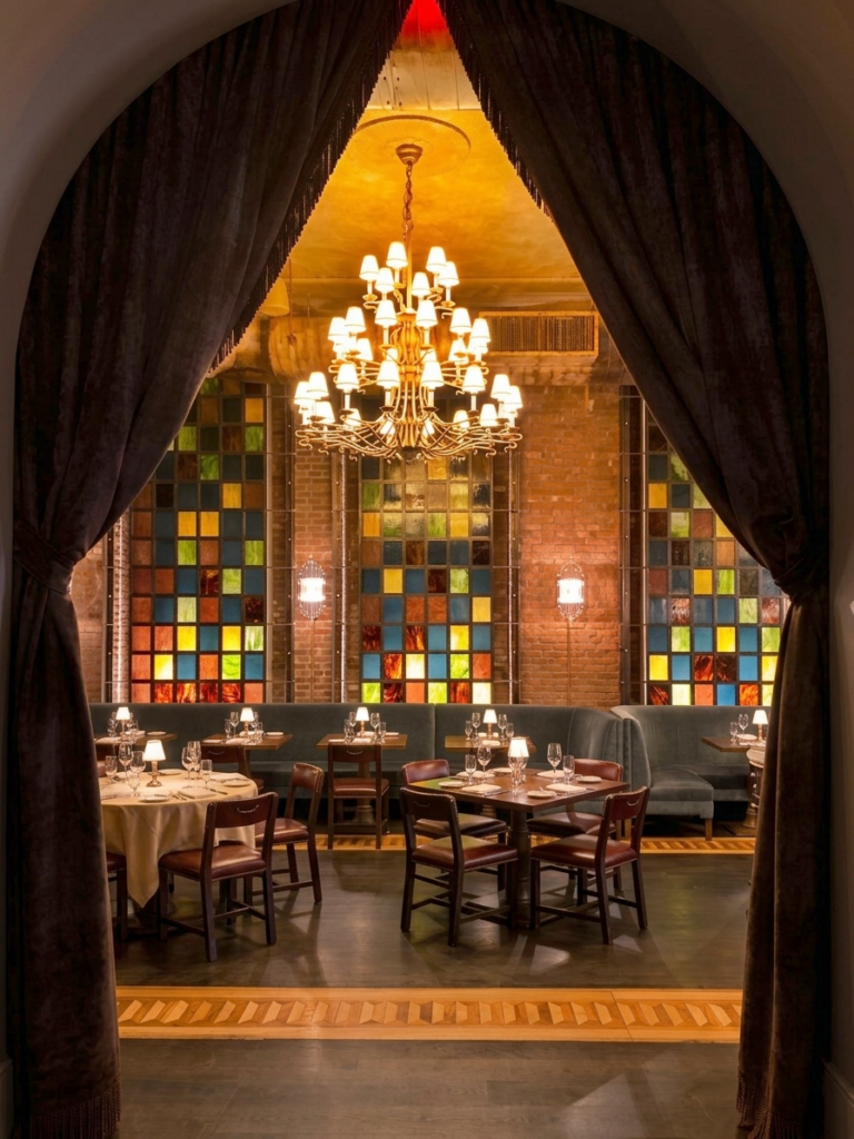 Dining room at The Beekman, New York framed by velvet drapery, with stained-glass windows, exposed brick walls, a multi-arm chandelier, blue banquettes, and dark wood tables.