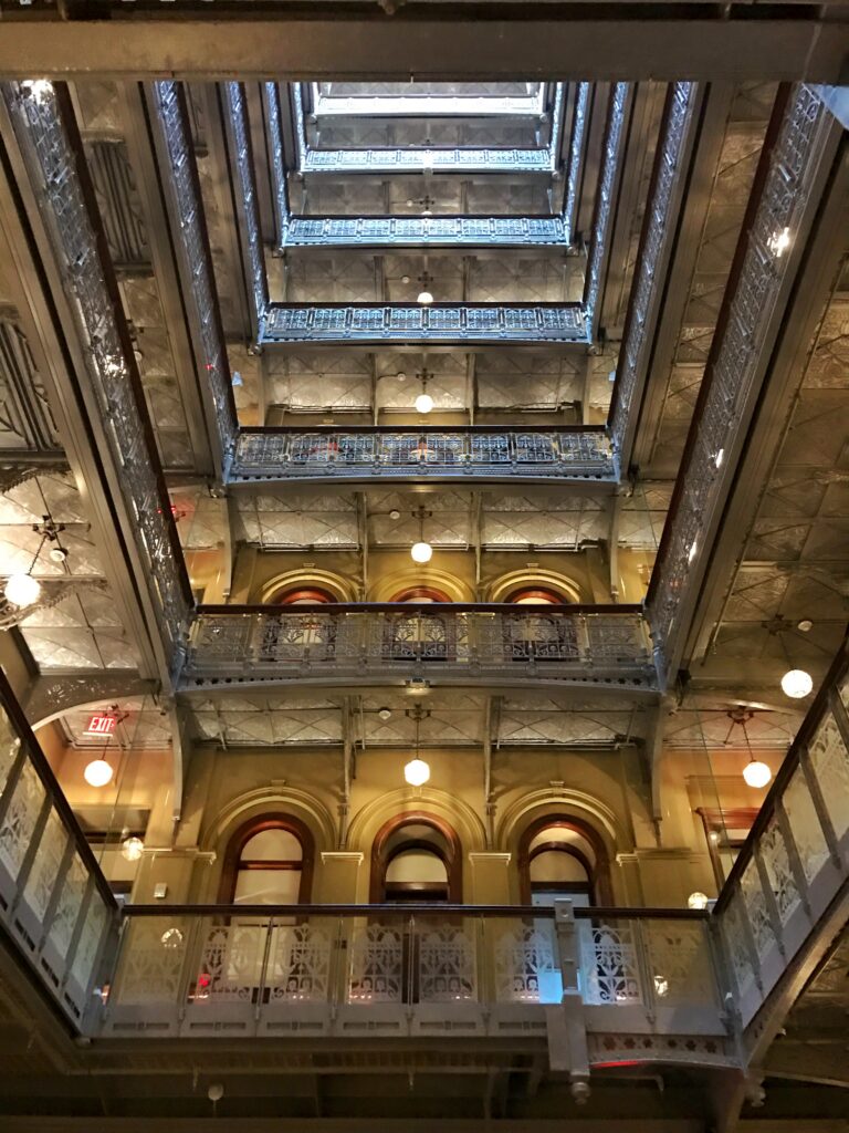 Central atrium at The Beekman Hotel, New York with stacked Victorian cast-iron balconies, arched windows, globe pendant lights, and a soaring historic interior framed by glass elevators.