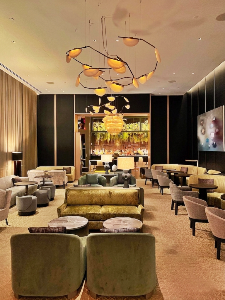 Lounge at The Ritz-Carlton New York, NoMad with olive velvet seating, curved banquettes, sculptural branch-like pendant lighting, and a softly glowing bar framed by floral arrangements.