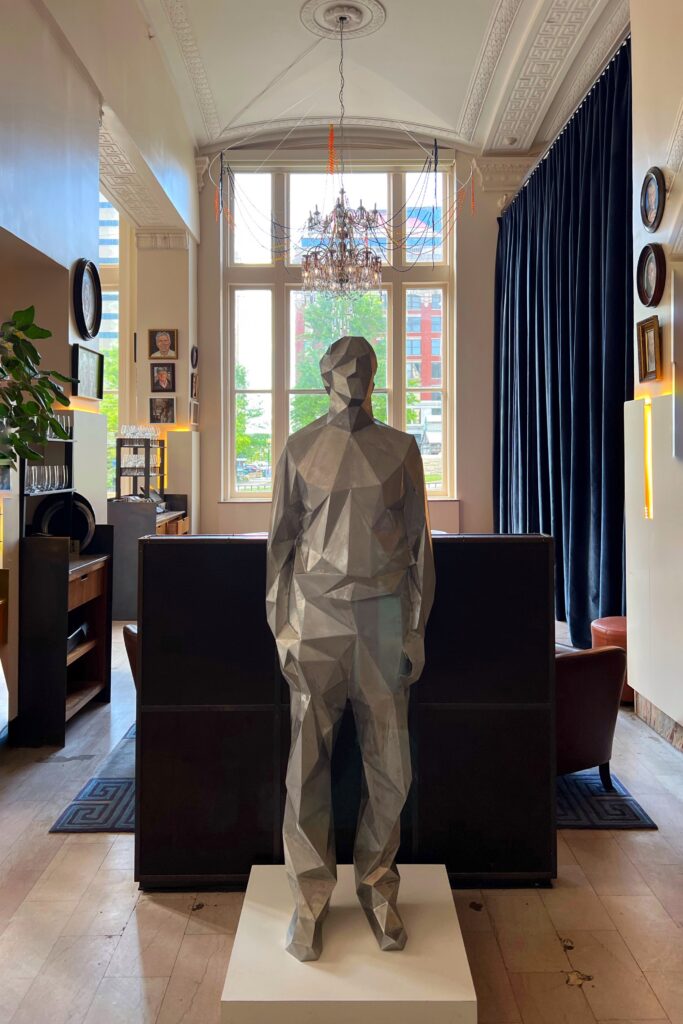 21c Museum Hotel lobby in Louisville, Kentucky with a faceted silver human sculpture and the iconic syringe chandelier hanging beneath ornate ceiling details, framed by tall windows and dark velvet curtains—an aesthetic hotel lobby and art museum.