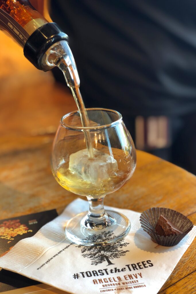 Bourbon tasting at Angel’s Envy Distillery in Louisville, Kentucky, with whiskey poured into a tulip glass over ice on a “#ToastTheTrees” napkin—an aesthetic stop on the Kentucky Bourbon Trail.