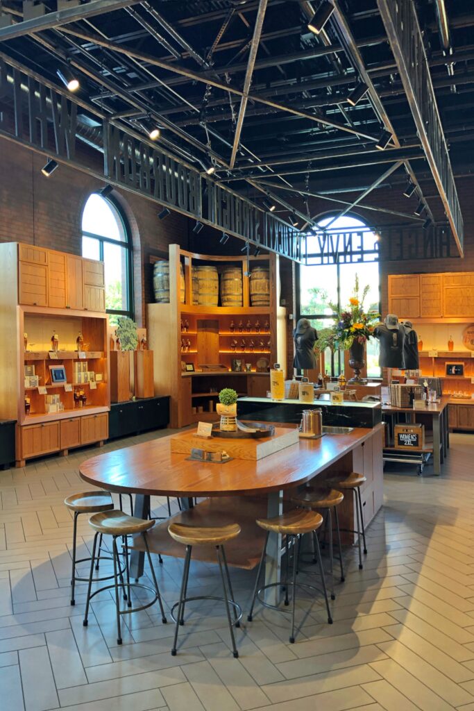 Angel's Envy Kentucky Bourbon Trail distillery gift shop and tasting room with arched factory windows, exposed steel ceiling, brick walls, and a central wood tasting table surrounded by bourbon displays and barrels.