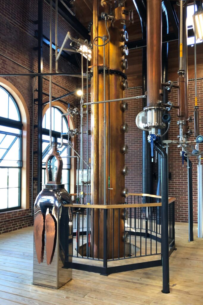 Angel’s Envy Distillery in Louisville, Kentucky featuring a tall copper still column with gauges and pipes, arched factory windows, exposed brick walls, and a reflective angel-wings sculpture—an aesthetic stop on the Kentucky Bourbon Trail.