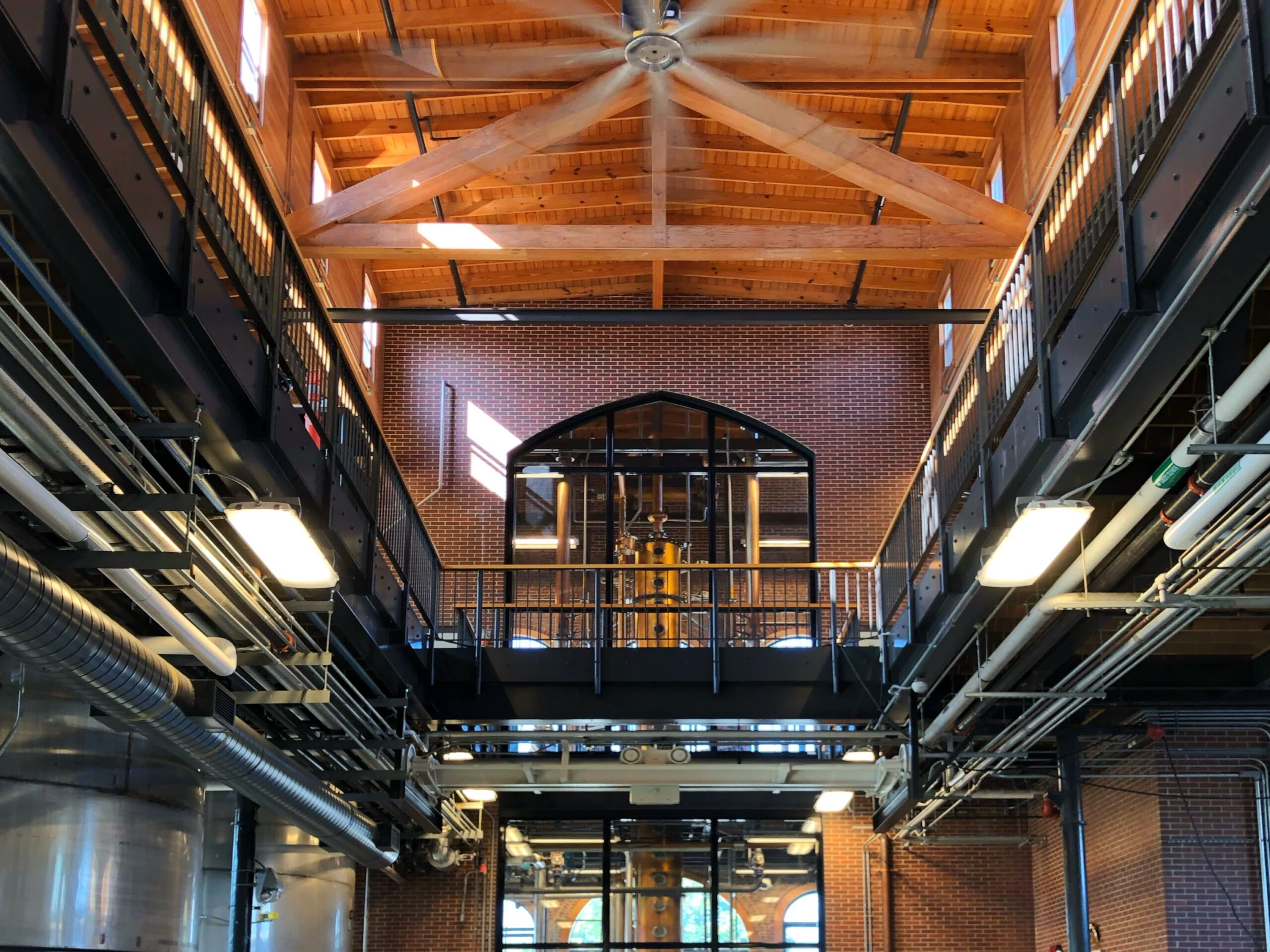 Aesthetic Bourbon Trail Travel Guide - Interior of Angel’s Envy Distillery in Louisville, Kentucky with exposed brick walls, steel catwalks, copper still behind an arched window, and a soaring wood-beam ceiling—an aesthetic stop on the Kentucky Bourbon Trail.