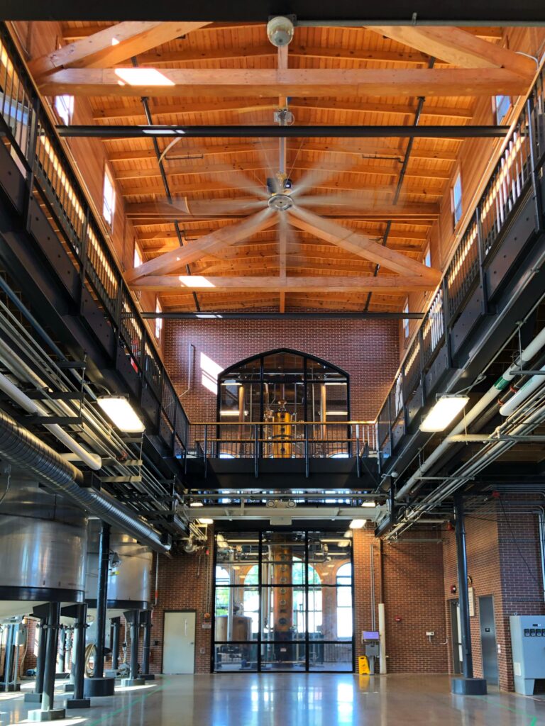 Angel’s Envy Distillery interior in Louisville, Kentucky featuring soaring timber trusses, exposed steel beams and pipes, tall factory windows, and a copper still visible behind glass—an aesthetic stop on the Kentucky Bourbon Trail.